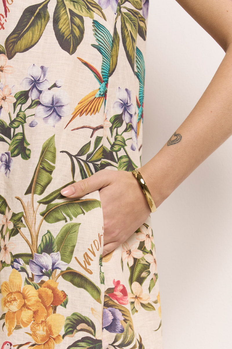 Eine Person in einem farbenfrohen Princess Goes Hollywood-Sommerkleid mit tropischem Aufdruck und bunten Blumen und Blättern steht mit der rechten Hand in der Tasche und zeigt ein goldenes Armband und ein kleines Herz-Tattoo auf ihrem Handgelenk vor einem schlichten Hintergrund.