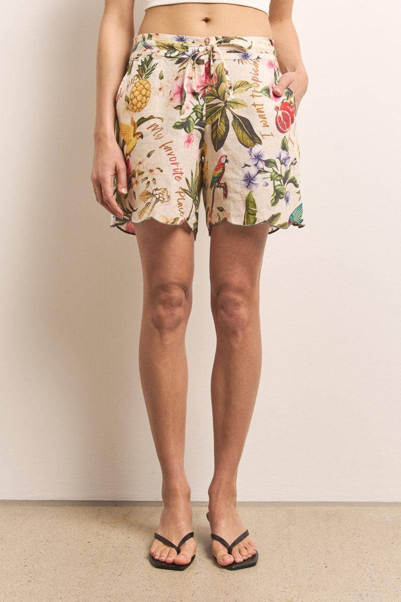 Eine Person steht vor einer schlichten Wand und trägt Princess Goes Hollywood Leinen-Shorts mit tropischen Motiven wie Vögel, Blumen und Ananas. Die Shorts haben einen gebundenen Bund und überschnittene Säume. Schwarze Flip-Flops vervollständigen den lässigen Look.