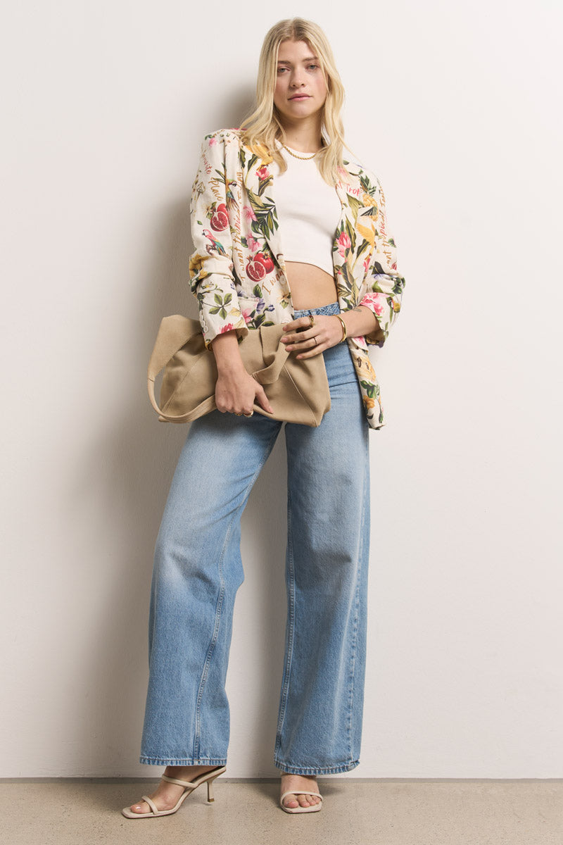 Eine Frau mit langen blonden Haaren steht selbstbewusst vor einer schlichten Wand. Sie trägt einen Leinenblazer mit tropischem Aufdruck von Princess Goes Hollywood über einem weißen, kurz geschnittenen Oberteil, hellblaue Jeans mit weitem Bein und beigefarbene Sandalen mit Absatz, in denen sie eine hellbraune Clutch hält.