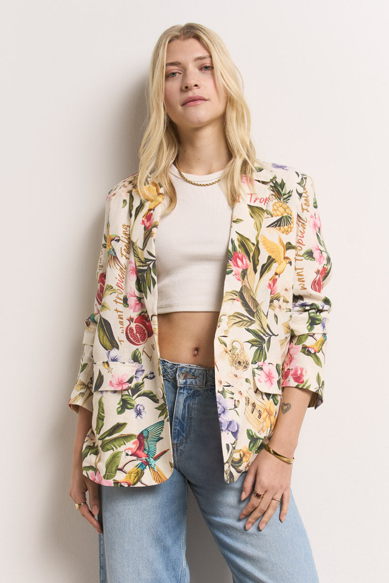 Eine Frau mit langen blonden Haaren trägt einen Princess Goes Hollywood Leinenblazer für Damen mit tropischem Aufdruck über einem weißen Crop-Top und hellblauen Jeans. Sie steht selbstbewusst vor einer schlichten weißen Wand und nimmt eine entspannte Haltung ein.