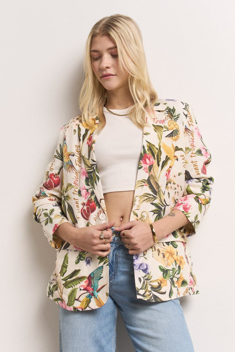 Eine junge Dame mit langen blonden Haaren trägt ein weißes Crop-Top, hellblaue Jeans und einen Princess Goes Hollywood Leinenblazer mit bunten Blumen und Vögeln im Tropenprint. Sie steht vor einer weißen Wand und schaut entspannt nach unten, die Hände halten ihren Blazer.