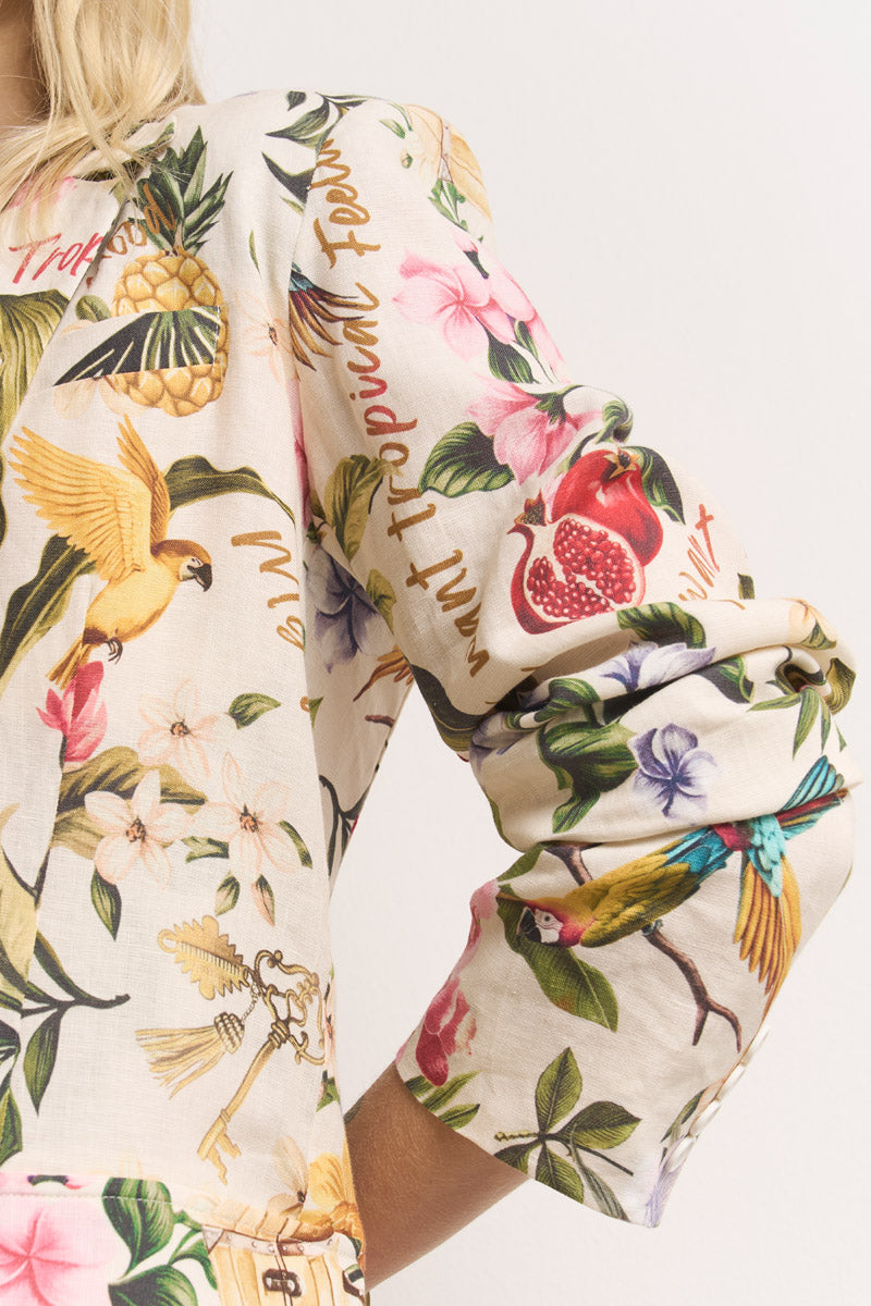 Nahaufnahme des Arms und des Oberkörpers einer Person, die ein cremefarbenes Princess Goes Hollywood-Tropical-Print-Shirt mit lebhaften Blumen, Papageien, Granatapfel, gelbem Vogel, tropischen Blättern und handschriftlichem Text trägt. Der Hintergrund ist einfarbig.
