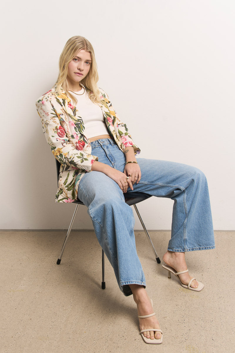 Eine Frau mit langen blonden Haaren sitzt auf einem schwarzen Stuhl vor einer schlichten Wand. Sie trägt einen Princess Goes Hollywood Damen Leinen Blazer, ein weißes Oberteil, hellblaue Jeans mit weitem Bein und Riemchensandalen mit Absatz und blickt sanft in die Kamera.