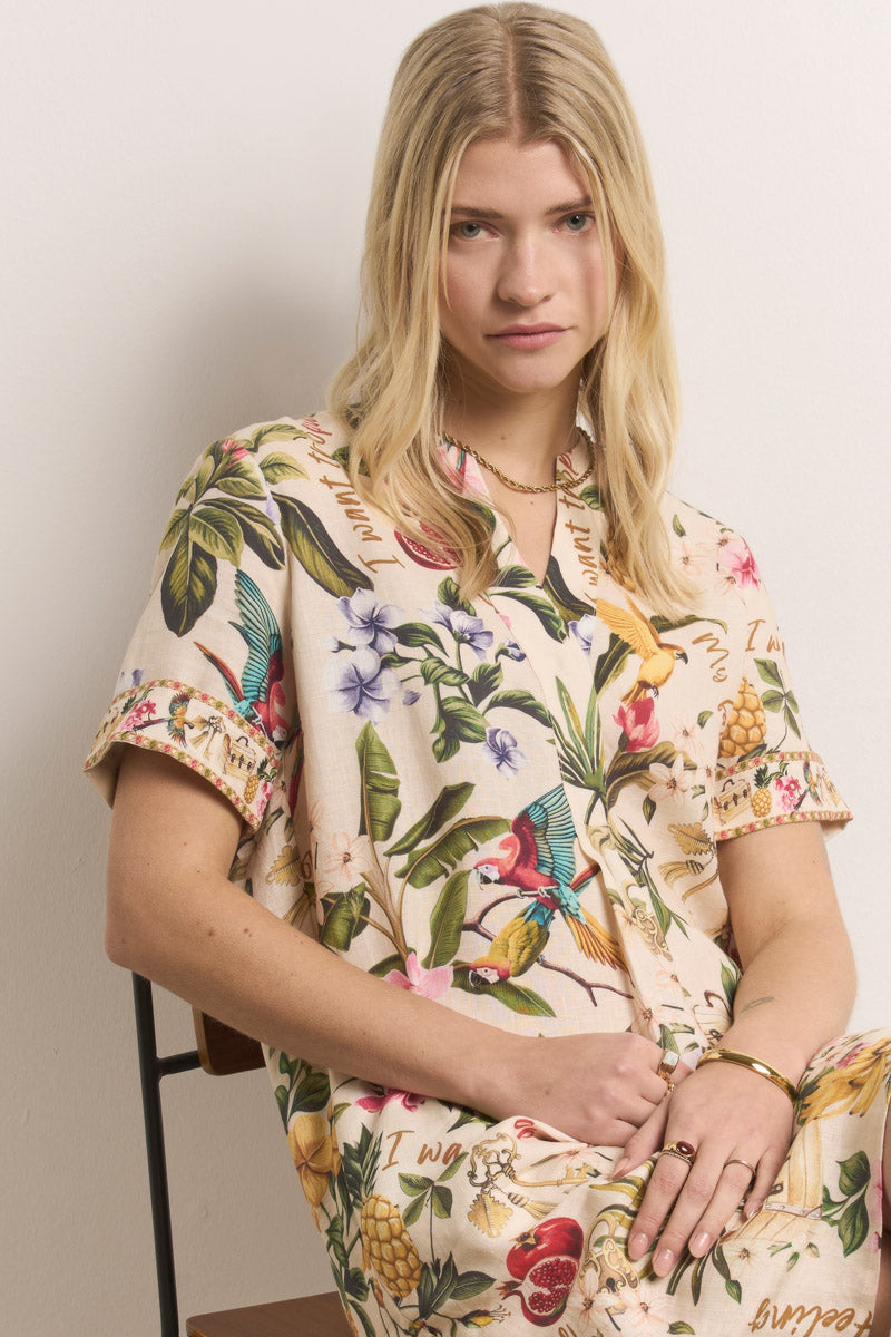 Eine junge Frau mit langen blonden Haaren sitzt auf einem Stuhl vor einer weißen Wand und trägt ein cremefarbenes Kurzkleid mit tropischen Motiven wie Vögel, Blumen und Früchte von Princess Goes Hollywood. Sie blickt neutral in die Kamera, die Hände ruhen in ihrem Schoß.