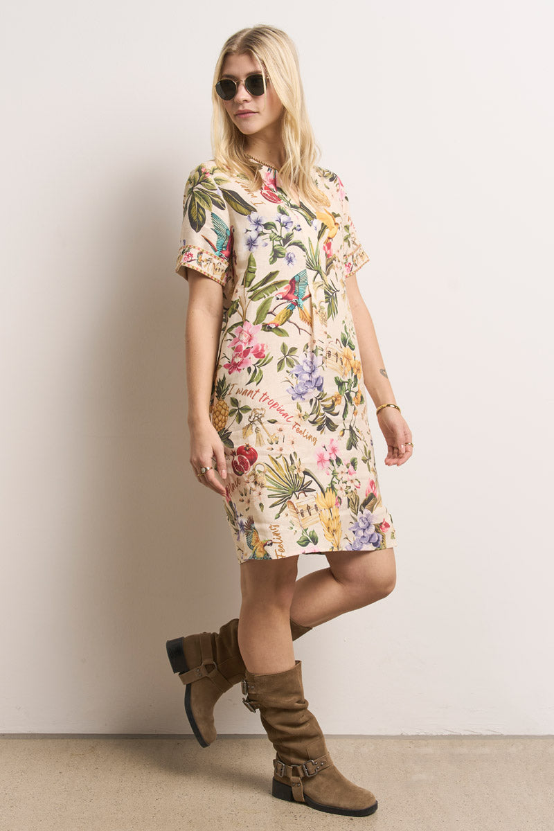 Eine Frau mit langen blonden Haaren trägt eine Sonnenbrille und ein Princess Goes Hollywood-Kurzkleid mit einem bunten tropischen Blumen- und Vogeldruck. Sie steht seitlich, nach links schauend, in braunen kniehohen Stiefeln vor einem schlichten Hintergrund.