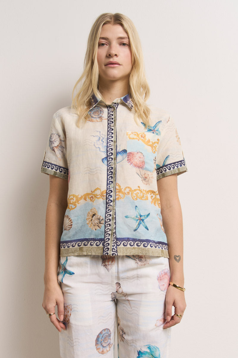 Eine Frau mit langen blonden Haaren trägt ein Princess Goes Hollywood-Leinenhemd mit Muscheldruck und eine passende Hose, beides kurzärmelig und mit maritimen Mustern in Blau, Orange und Beige. Sie steht vor einem einfarbig weißen Hintergrund und blickt geradeaus.