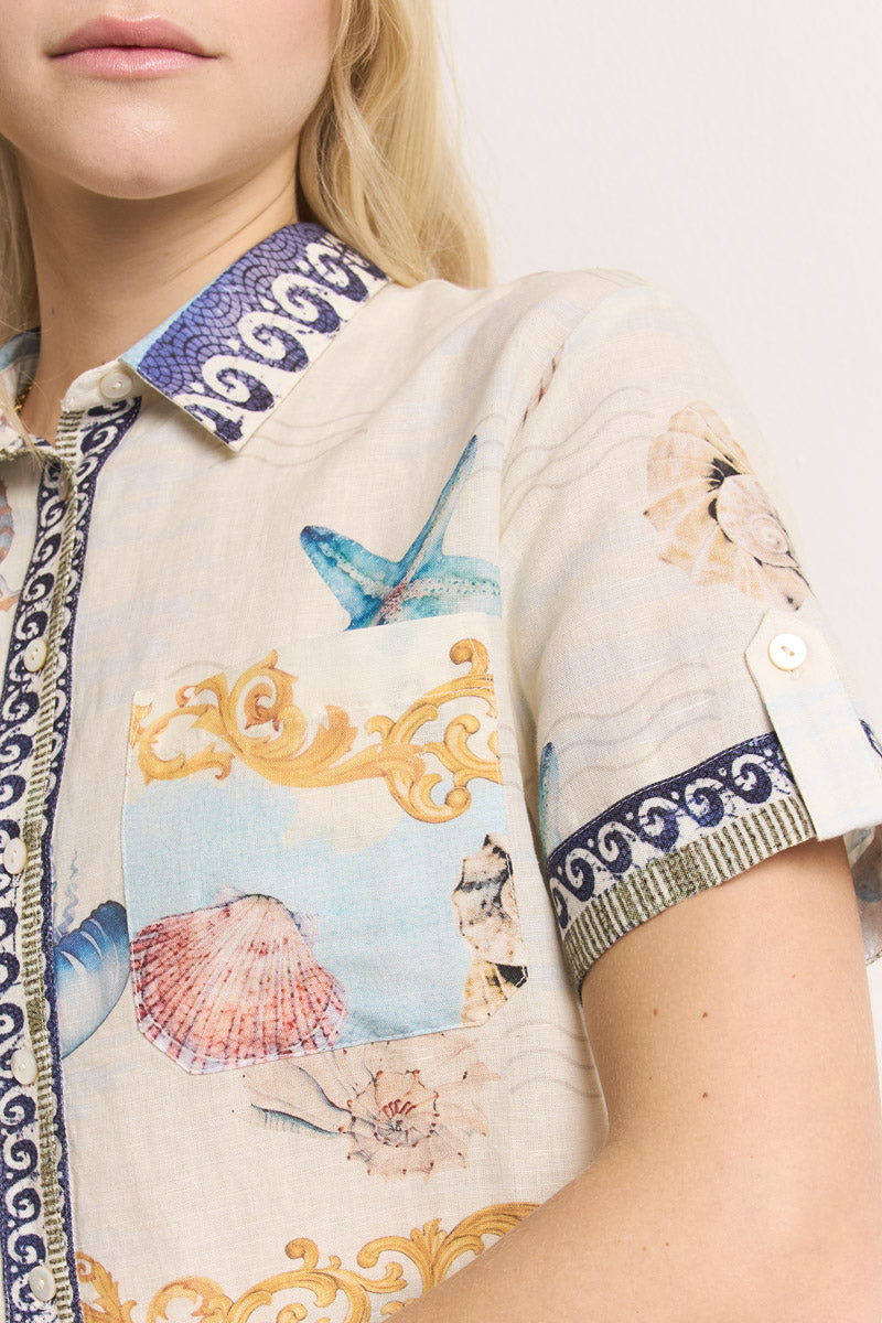 Eine Frau trägt eine beigefarbene Princess Goes Hollywood Leinen-Hemdbluse mit kurzen Ärmeln, mit blau-goldenem Muschel-Print, gemustertem Kragen und Ärmelbesatz, einer Brusttasche mit Muschelmuster, cremefarbenen Knöpfen, und nur ihr unteres Gesicht ist sichtbar.