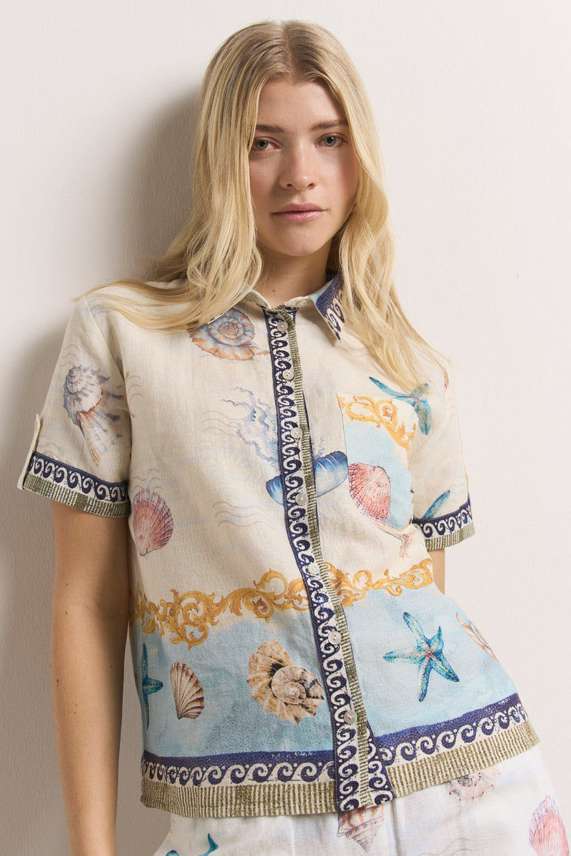 Eine blonde Frau mit offenem Haar steht vor einer hellen Wand und trägt eine Princess Goes Hollywood-Leinenbluse mit kurzen Ärmeln. Die Bluse ist mit Muscheln, Seesternen und Wellen in Blau, Beige und Gold bedruckt.