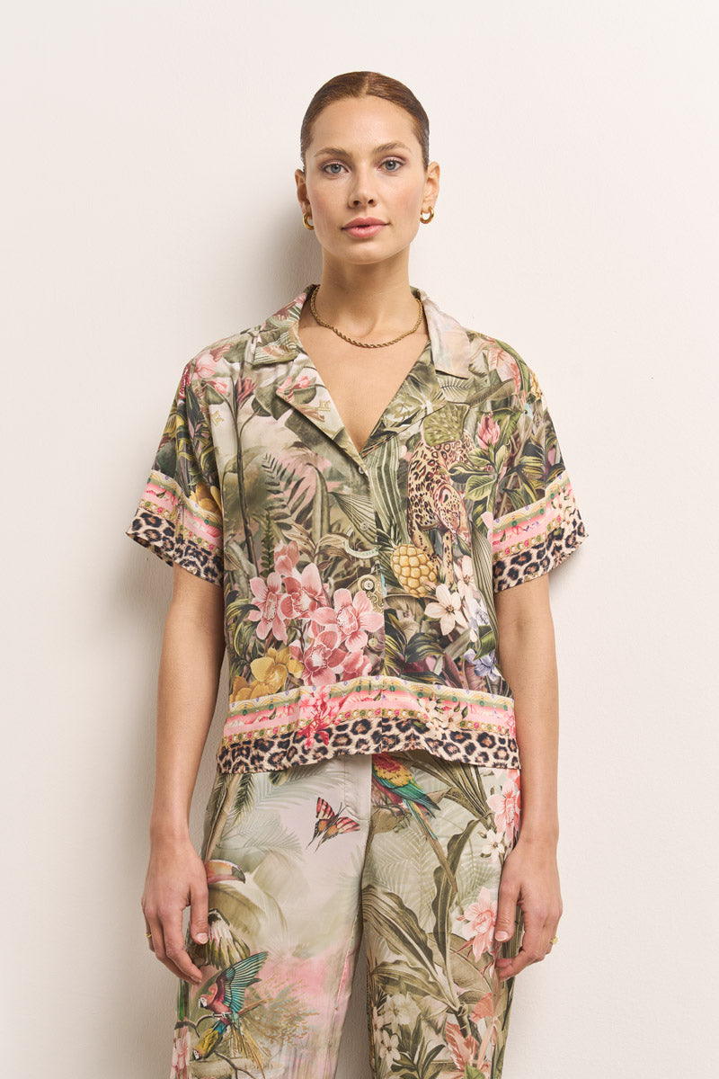 Eine Frau mit zurückgekämmtem Haar, goldenen Ohrringen und einer goldenen Halskette trägt eine kurze Princess Goes Hollywood-Hemdbluse und eine dazu passende Hose mit einem bunten Dschungel-Leopardenmuster, einem gekerbten Kragen und tropischen Motiven vor einem hellen Hintergrund.