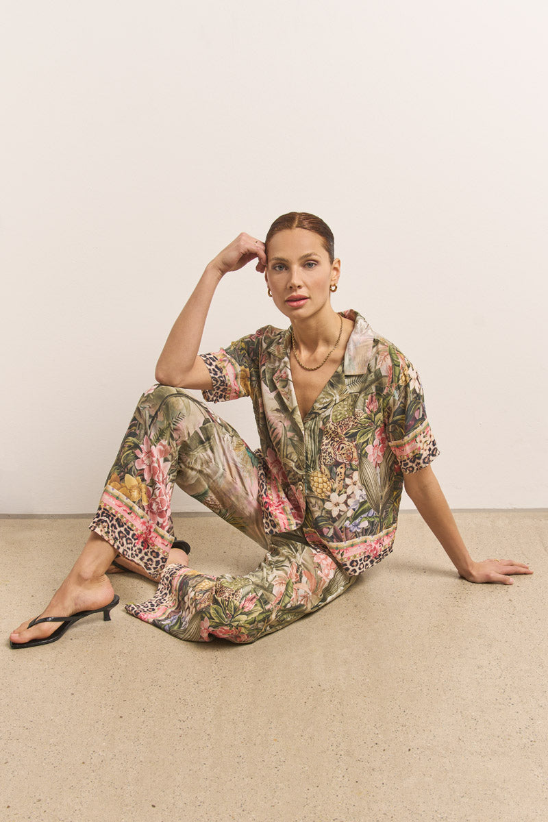 Eine Frau mit hochgestecktem Haar sitzt auf dem Boden und trägt die kurze Hemdbluse von Princess Goes Hollywood und eine kurze Hose in einem leuchtenden Dschungel-Leo-Print, gepaart mit schwarzen hochhackigen Sandalen. Ein schlichter weißer Hintergrund unterstreicht ihren Reverskragen-Stil.