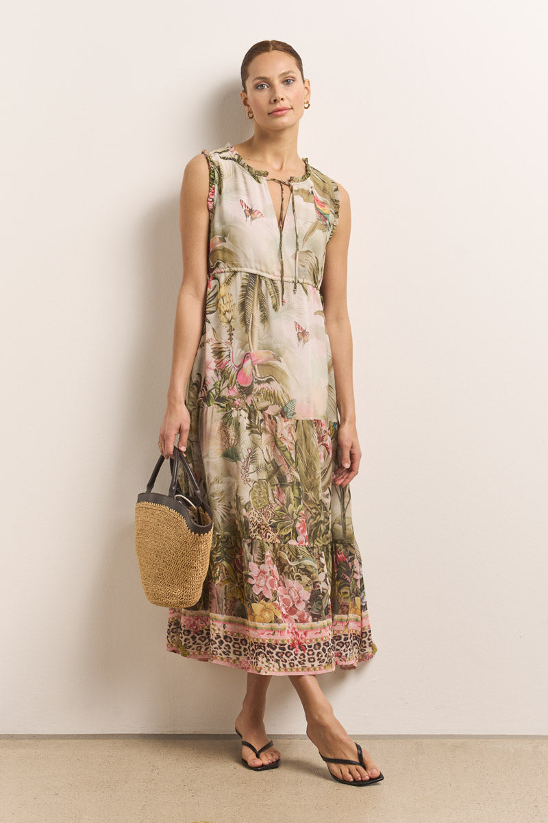 Eine Frau steht vor einer weißen Wand und trägt ein ärmelloses, knöchellanges Kleid mit Dschungel-Leo-Print in Rosa und Grün von Princess Goes Hollywood. Sie trägt eine geflochtene Strohhandtasche, schwarze Flip-Flops und goldene Ohrringe, ihr Haar ist hochgesteckt.