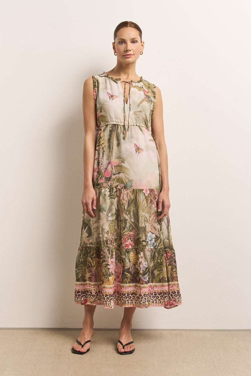 Eine Frau mit hochgesteckten rötlichen Haaren trägt ein tropisch-blumiges Princess Goes Hollywood-Midikleid in sanften Farbtönen, das eine lockere Passform, einen V-Ausschnitt mit Bindeband und einen gestuften Rock aufweist. Sie steht in schwarzen Flip-Flops vor einer schlichten Wand.