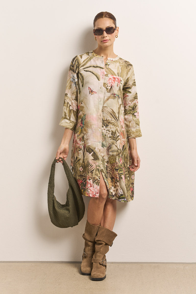 Eine Frau steht an einer schlichten Wand und trägt ein Dschungel-Leo-Print langärmliges Leinen Blusenkleid von Princess Goes Hollywood, braune Schlabberstiefel, eine dunkle Sonnenbrille, goldene Ohrringe und eine olivgrüne strukturierte Handtasche. Ihr Haar ist zurückgesteckt und sie lächelt sanft.