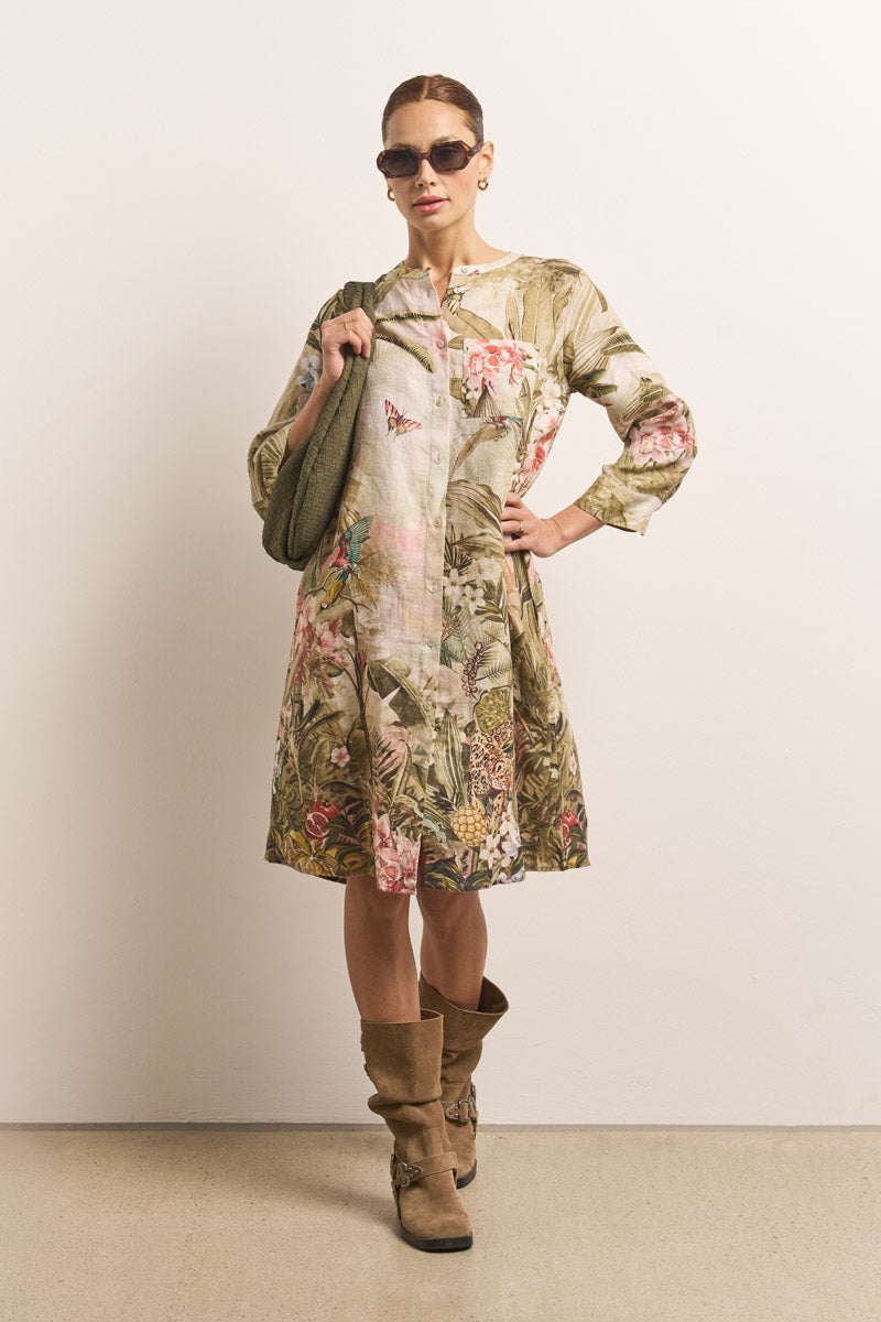 Eine Frau posiert vor einer schlichten hellen Wand in einem knielangen Princess Goes Hollywood Damenkleid mit erdigen Blumenmustern, braunen kniehohen Stiefeln, Sonnenbrille, einer olivgrünen Stricktasche und hochgestecktem Haar.