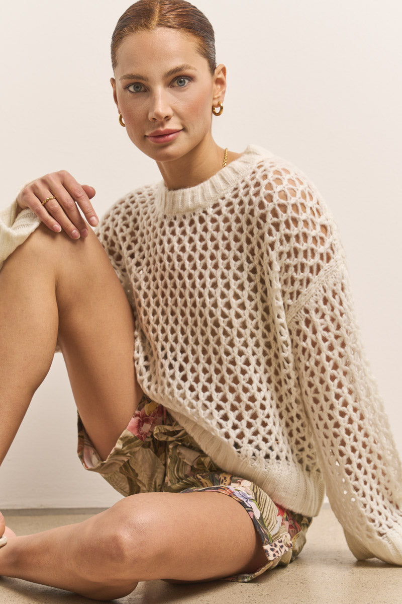 Eine Frau mit hochgestecktem Haar und goldenen Reifen sitzt an einer hellen Wand und lächelt sanft. Sie trägt einen cremefarbenen, offenen Strickpullover und dazu eine Princess Goes Hollywood Jungle-Leo-Print Damenhose, ein Knie dicht vor der Brust angewinkelt.