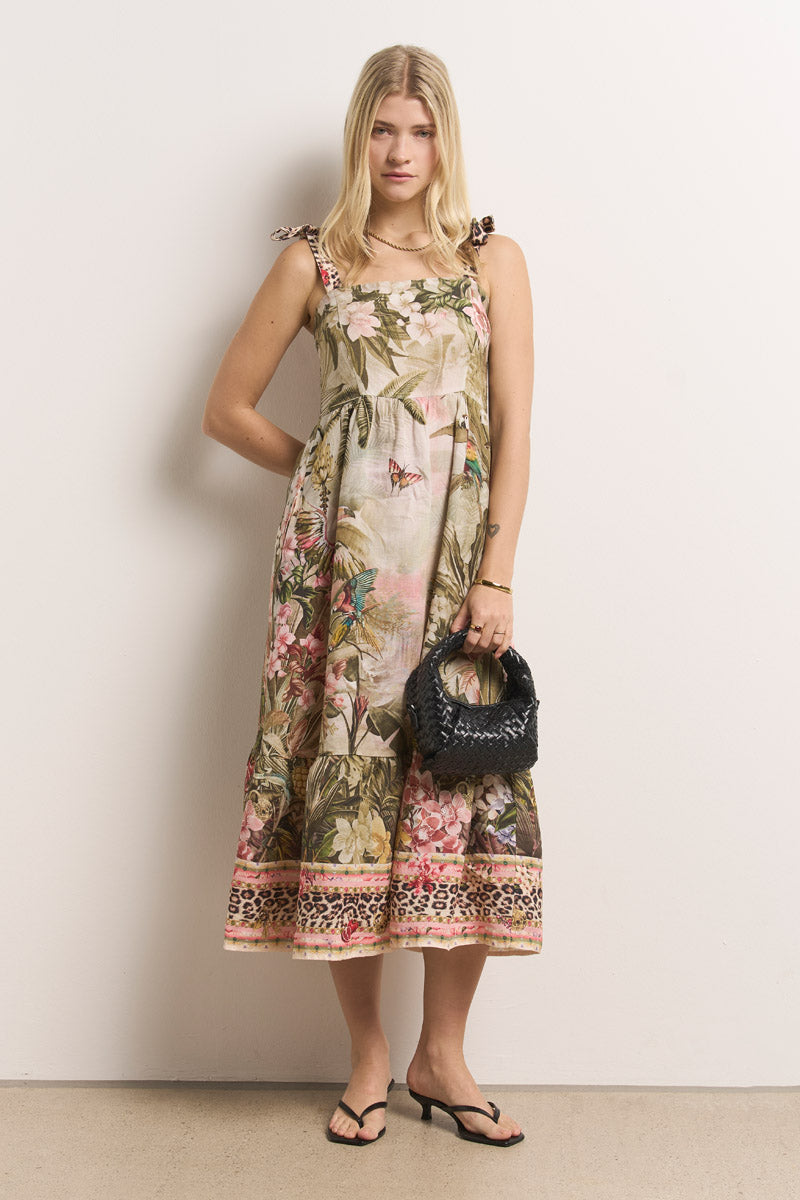 Eine Frau mit langen blonden Haaren trägt ein Princess Goes Hollywood Leinen-Midikleid mit Bindebändern und einem volantförmigen Saum. Sie hält eine schwarze, strukturierte Handtasche in der Hand, trägt schwarze Flip-Flop-Sandalen, steht an einer hellen Wand und schaut in die Kamera.