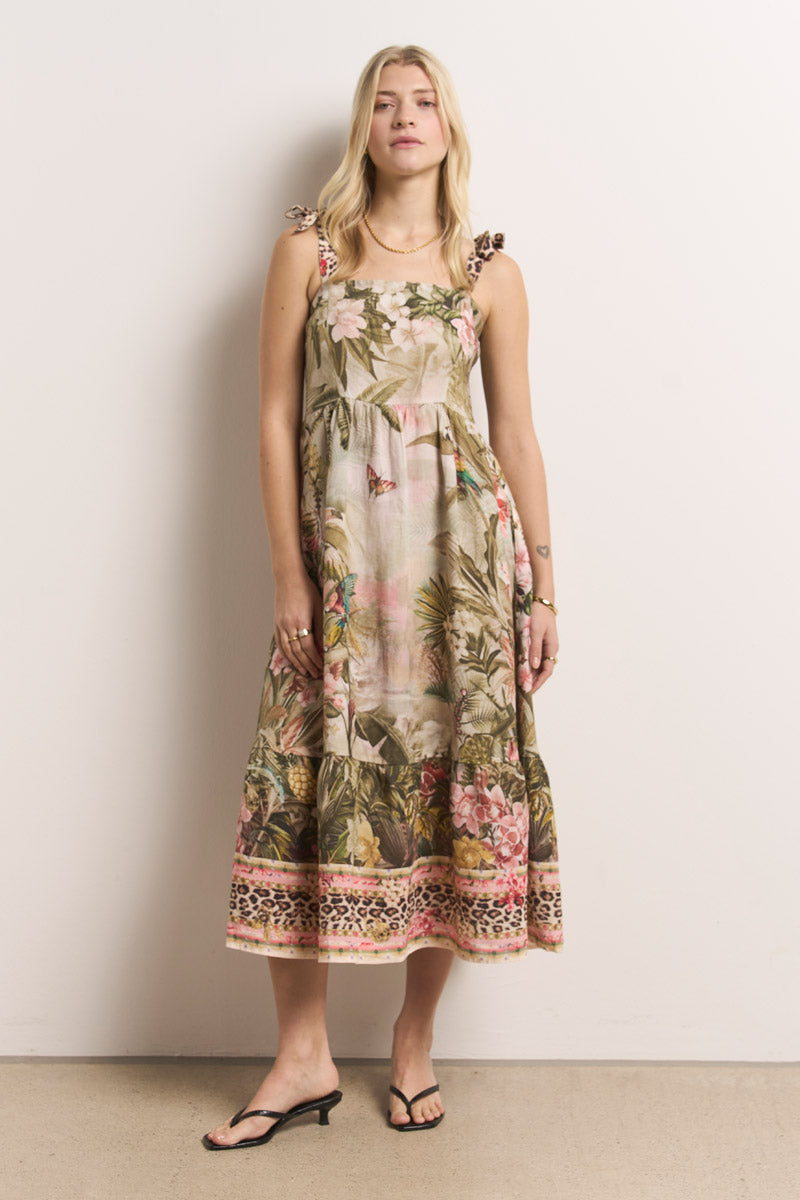 Eine Frau mit langen blonden Haaren trägt ein ärmelloses Princess Goes Hollywood Leinen-Midikleid mit tropischem Blumen- und Dschungel-Leo-Print, Schleifen an den Schultern, einem bunten Saum und schwarzen Sandalen und steht vor einer gebrochen weißen Wand.