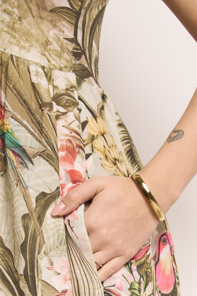 Jemand trägt ein Princess Goes Hollywood Leinen Midikleid mit einem lebhaften Dschungel-Leo-Print aus grünen Blättern und bunten Vögeln, die Hand in der Tasche. Dazu trägt sie ein goldenes Armband und ein kleines Unterarmtattoo auf hellem, schlichtem Hintergrund.