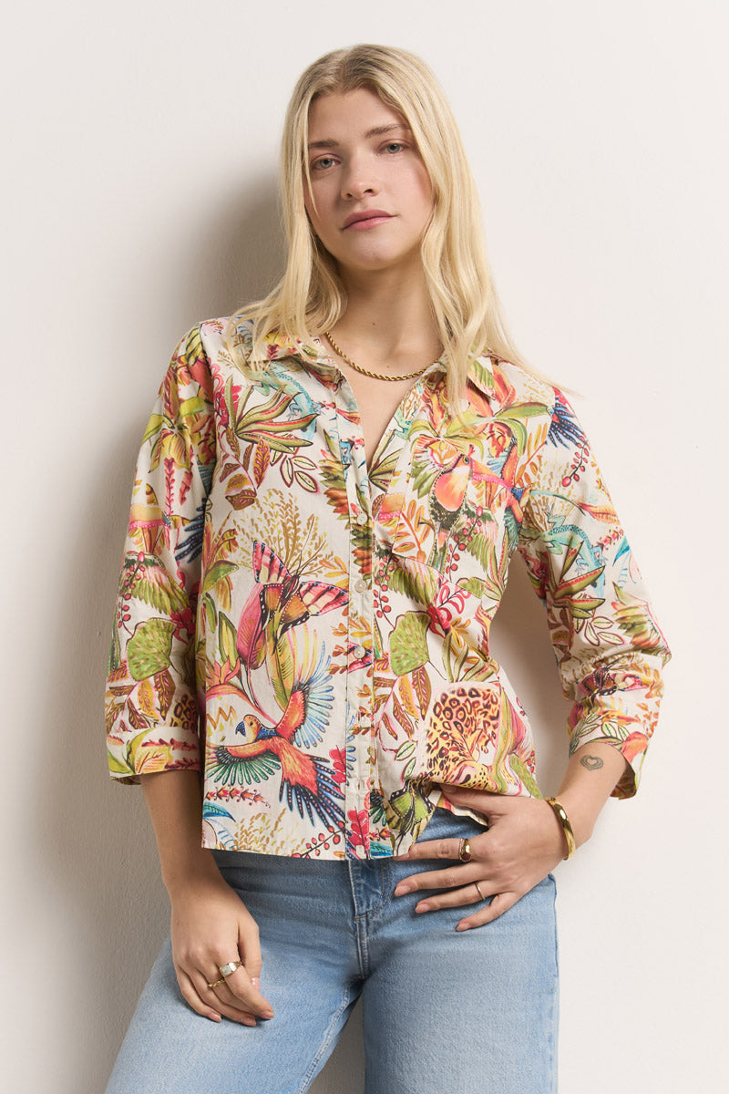 Eine junge Frau mit langen blonden Haaren trägt eine leuchtende Princess Goes Hollywood Hemdbluse aus Baumwolle mit bunten Vögeln und Blättern. Sie steht vor einer schlichten hellen Wand, eine Hand in der Jeanstasche, und blickt selbstbewusst in die Kamera.