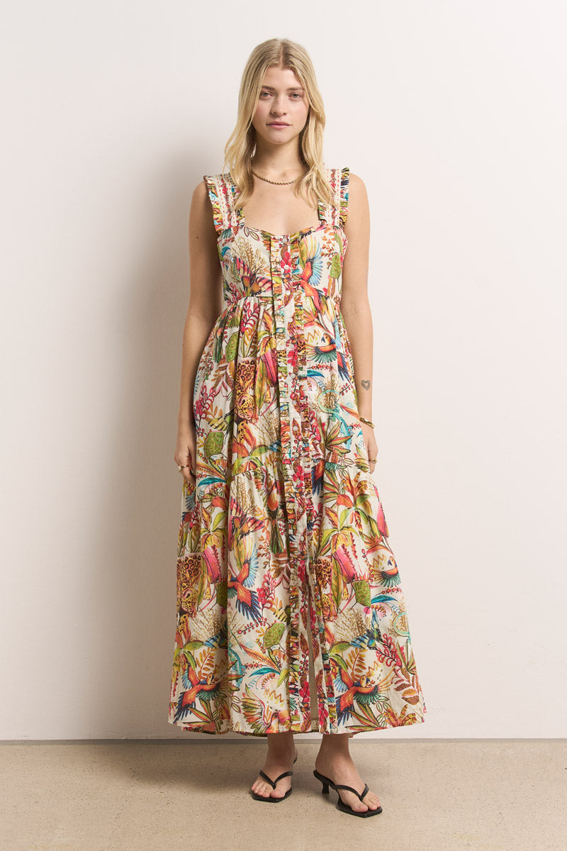 Eine Frau mit blondem Haar trägt ein farbenfrohes Princess Goes Hollywood Maxikleid aus Baumwolle mit Dschungel-Print, gerüschten Trägern und einem Stufenrock, gepaart mit Sandalen und minimalem Schmuck für eine mühelose Sommeratmosphäre vor einer schlichten weißen Wand.