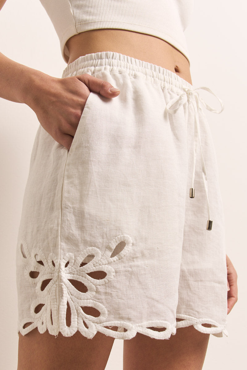 Nahaufnahme einer Person, die die weißen Leinenshorts Princess Goes Hollywood" mit Stickereiausschnitt, überschnittenem Saum, elastischem Bund mit Kordelzug und einer Hand in einer Tasche neben einem weißen gerippten Crop-Top trägt.