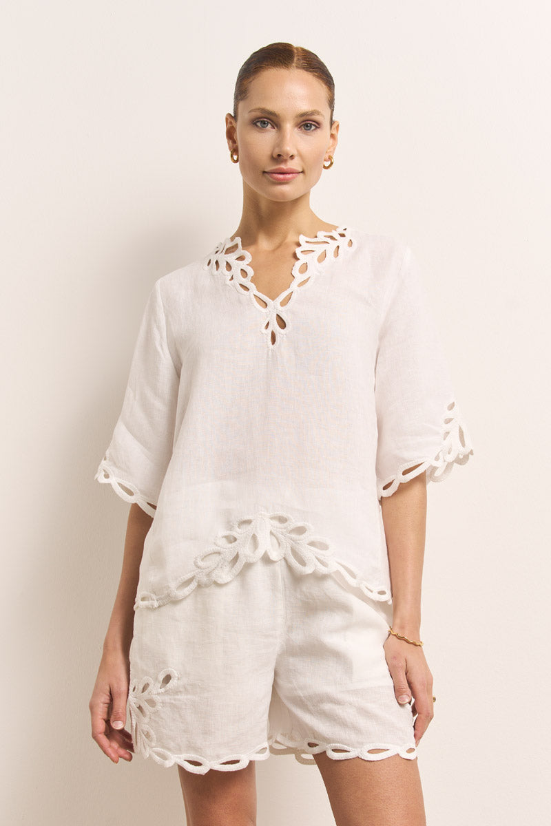 Eine Frau steht vor einem schlichten hellen Hintergrund und trägt eine weiße Leinenbluse und passende Shorts von Princess Goes Hollywood. Beide Teile haben einen elastischen Bund, Seitentaschen, Wellenkanten und ausgeschnittene Blumendetails. Dazu trägt sie goldene Reifohrringe und einen Armreif.