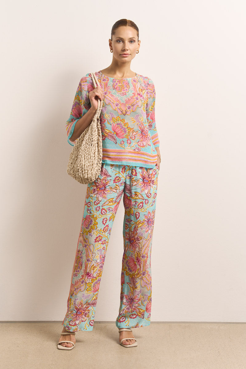 Eine Frau posiert vor einer schlichten Wand in einem lockeren, farbenfrohen Blumenprint-Top und einer passenden fließenden Hose mit elastischem Bund in Pink, Aqua und Gelb von Princess Goes Hollywood. Dazu trägt sie eine beigefarbene Umhängetasche aus Netzstoff, Reifohrringe und Sandalen.