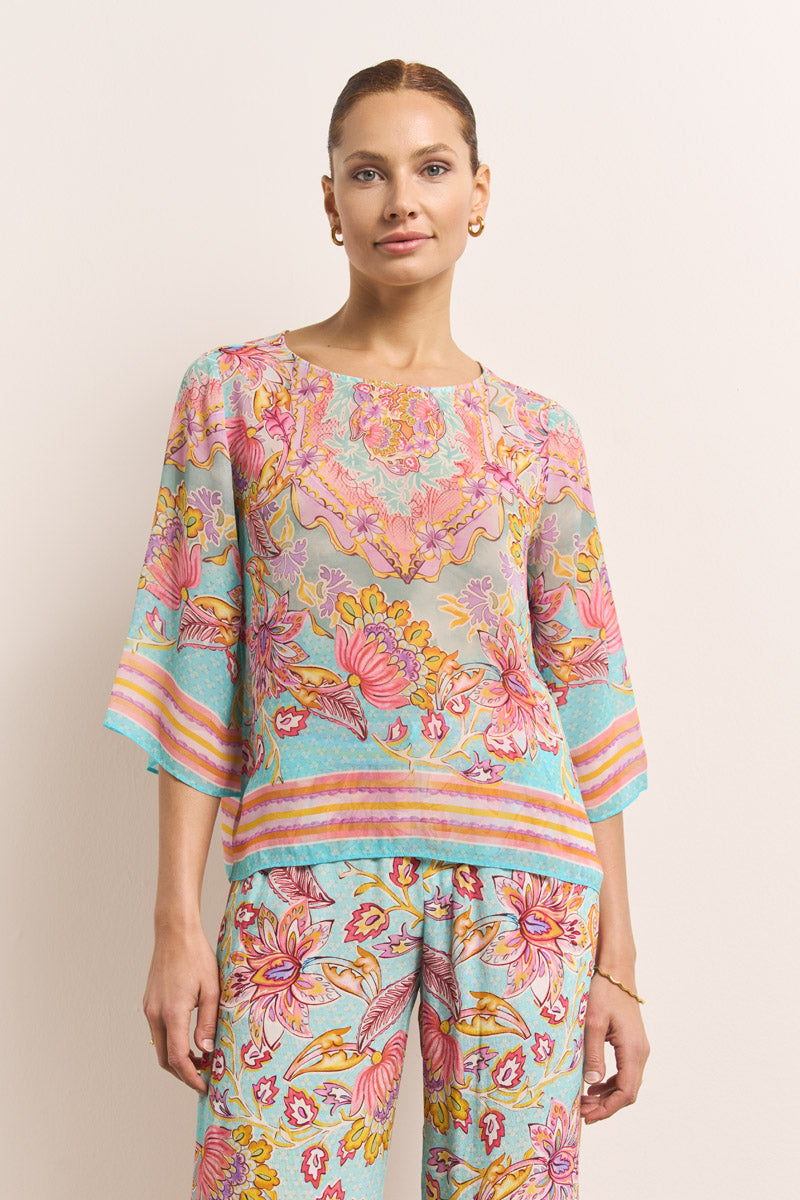 Eine Frau mit hellbraunen Haaren trägt eine farbenfrohe Blumenprint-Bluse und eine fließende Hose mit elastischem Bund in Pink, Gelb, Aqua und Orange von Princess Goes Hollywood. Sie steht vor einem schlichten off-white Hintergrund.