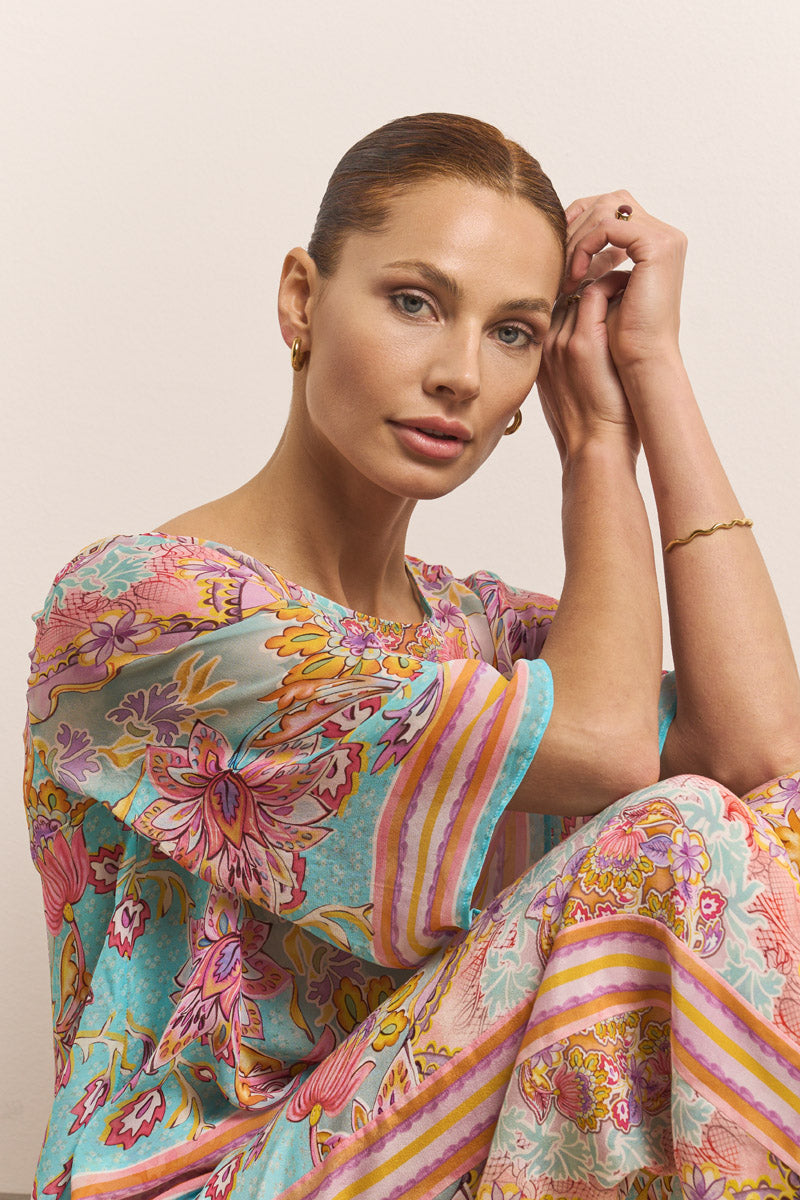 Eine hellhäutige Frau mit zurückgebundenem kastanienbraunem Haar blickt sanft in die Kamera. Sie trägt ein leuchtendes türkis-, pink- und goldfarbenes Blumenprint-Kleid von Princess Goes Hollywood, dazu goldene Creolen und ein zartes Armband vor einem neutralen Hintergrund.