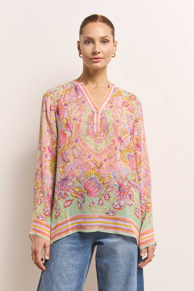 Eine Frau steht vor einer schlichten, hellen Wand und trägt eine Princess Goes Hollywood-Tunika mit V-Ausschnitt und leuchtendem rosa, grünen und gelben Blumenmuster. Dazu trägt sie eine hellblaue Jeans, goldene Ohrringe und hat ihr Haar ordentlich zurückgekämmt.