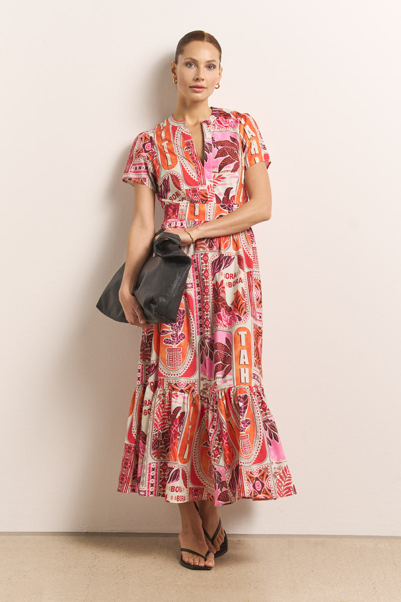 Eine Frau mit zurückgekämmtem Haar posiert an einer schlichten Wand und trägt ein leuchtendes Princess Goes Hollywood Maxikleid aus Baumwollpopeline mit auffälligem Tahiti-Print in Rot, Orange und Pink. Sie trägt eine große schwarze Tasche und schwarze Sandalen.