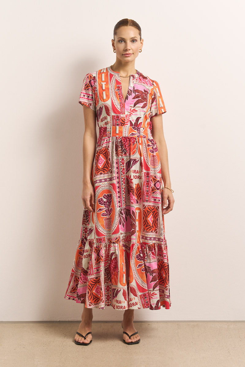 Eine Frau steht vor einer schlichten Wand und trägt ein Princess Goes Hollywood Baumwollpopeline Maxikleid mit kurzen Ärmeln, einem gestuften Rock und leuchtendem Tahiti-Print in Pink, Rot, Orange und Creme. Dazu trägt sie einfache Sandalen und kleine Ohrringe.