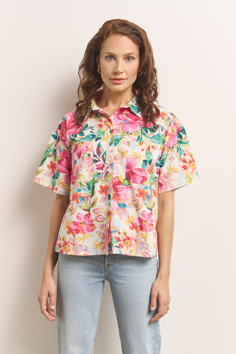 Eine Frau mit gewelltem kastanienbraunem Haar modelliert eine Princess Goes Hollywood bunte Hemdbluse aus Baumwolle mit Blumenprint in Pink, Grün und Gelb. Sie steht frontal, Hände an den Seiten, kombiniert dazu eine hellblaue Jeans vor hellem Hintergrund.