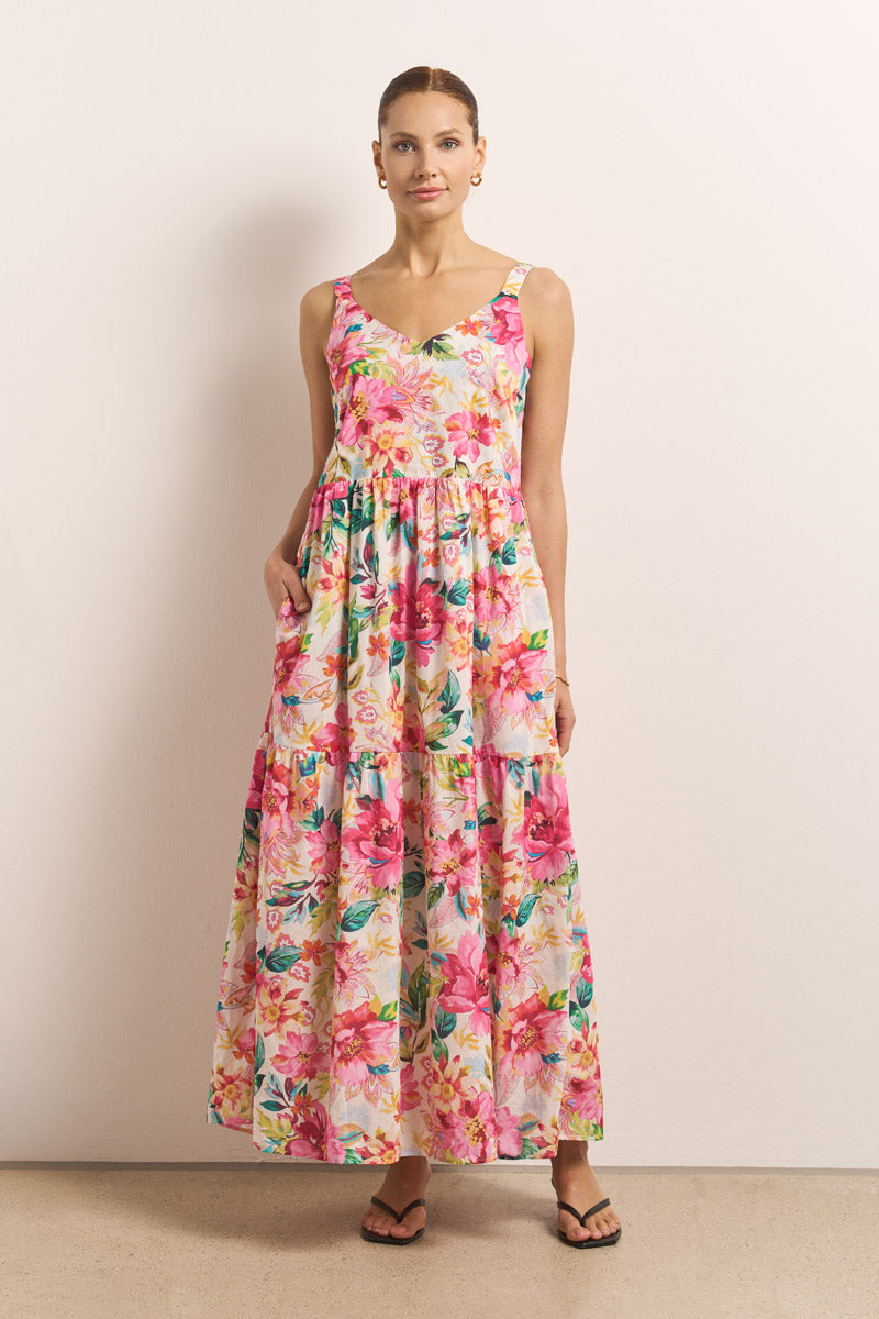 Eine Frau steht vor einer hellen Wand und trägt ein ärmelloses Princess Goes Hollywood-Baumwollkleid mit einem leuchtenden rosa, roten und gelben Blumendruck. Ihr Haar ist hochgesteckt, und sie trägt Ohrringe und schwarze Sandalen zu den Taschen ihres Kleides.