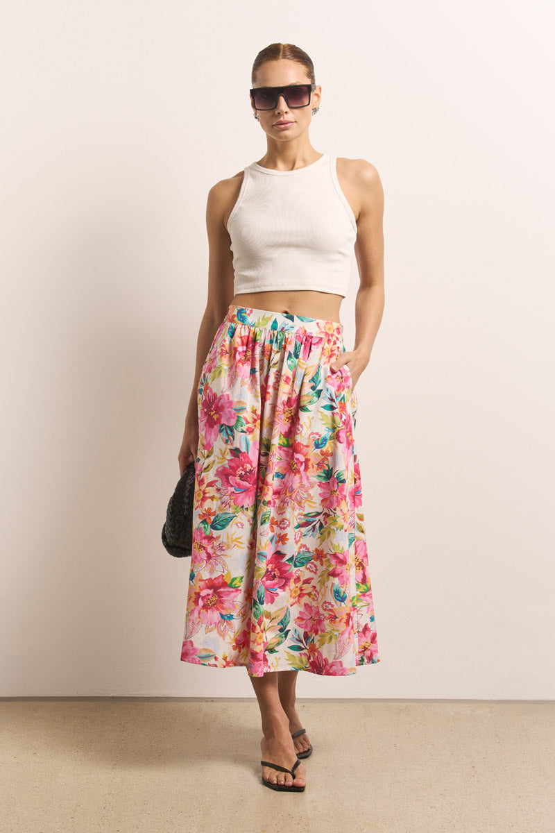 Eine Frau trägt ein weißes, ärmelloses Crop-Top, einen bunten Blumen-Midirock und eine rechteckige Okkia Sonnenbrille mit UV 400 Schutz. Sie hält eine schwarze Clutch, trägt schwarze Sandalen und hat zurückgebundene Haare.