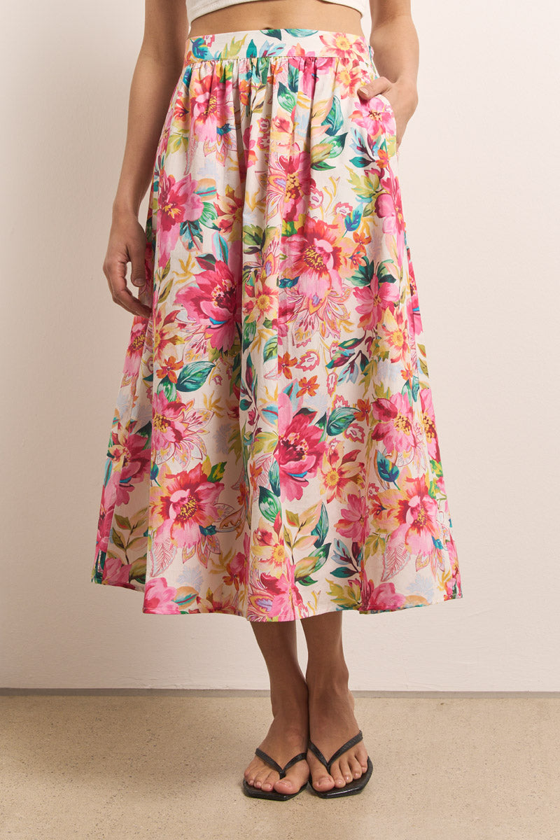 Jemand trägt einen Princess Goes Hollywood Midirock aus Baumwolle mit hoher Taille und Plisseefalten, der mit einem Blumenmuster in Pink, Orange, Gelb und Grün bedruckt ist und Taschen hat. Gestylt mit schwarzen Flip-Flops und einem leichten Top stehen sie vor einer schlichten Wand.