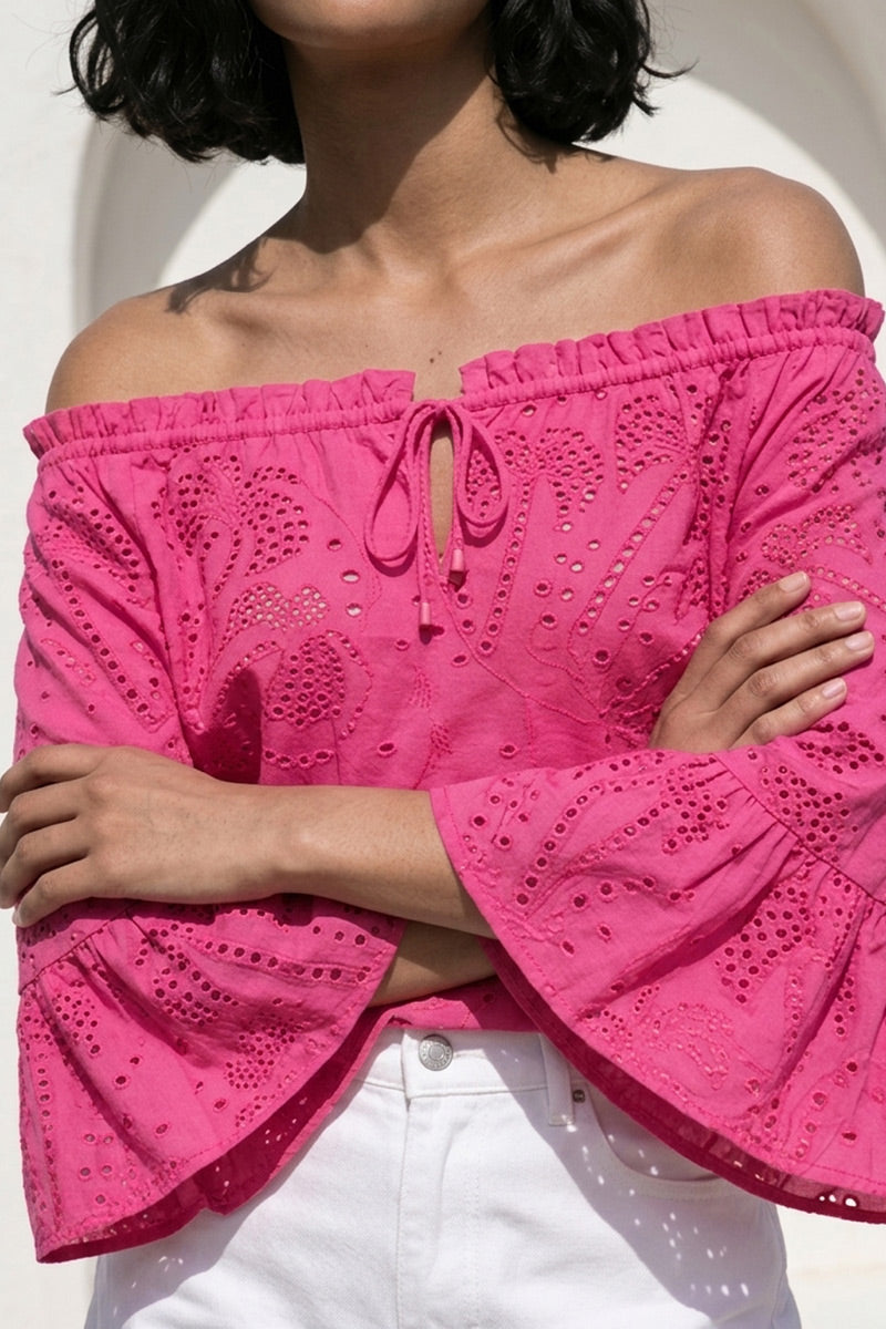 Eine Person mit kurzen dunklen Haaren trägt eine leuchtend pinke schulterfreie Carmenbluse mit Lochstickerei, Volantärmeln und einer Krawatte vorne von Princess Goes Hollywood, gepaart mit hochgekrempelten weißen Jeans. Die Arme sind vor einem sanft verschwommenen Hintergrund gekreuzt.