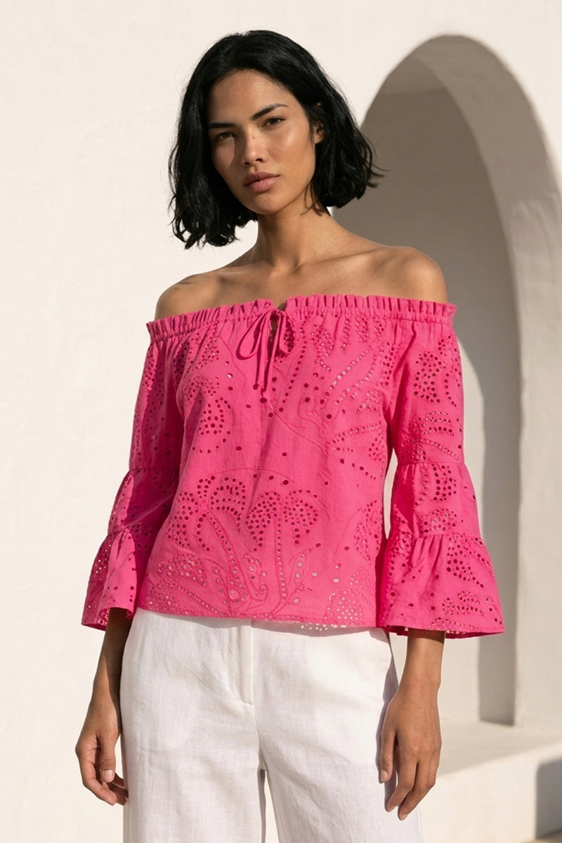 Eine Frau mit mittlerem Hautton und kurzen schwarzen Haaren trägt eine leuchtend pinke Princess Goes Hollywood Carmenbluse mit Lochstickerei und 3/4-Ärmeln, gestylt mit einer weißen Hose, und steht vor einer weißen Wand und einer gewölbten Tür.