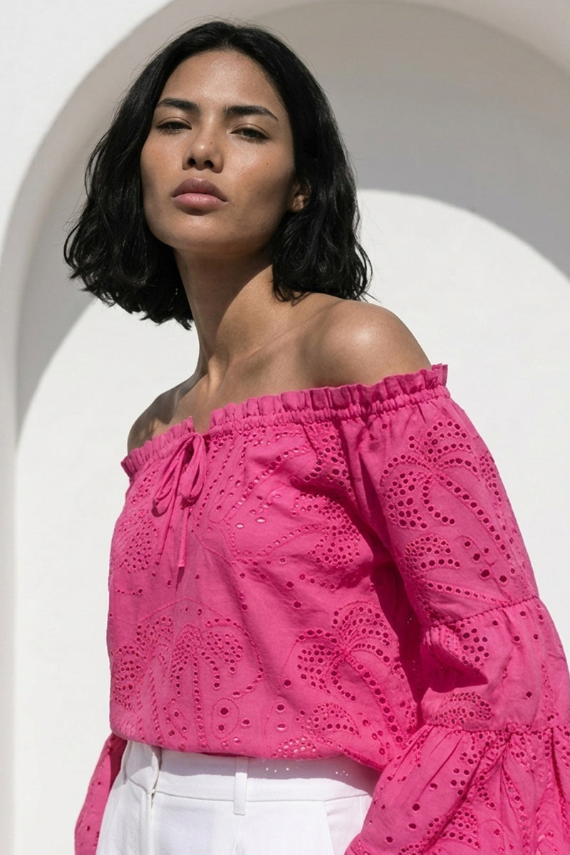 Eine Frau mit mittlerer Hautfarbe und schulterlangem dunklem Haar trägt eine leuchtend pinke Carmenbluse mit Volantärmel und Lochstickerei von Princess Goes Hollywood, gepaart mit einer weißen Hose mit hoher Taille. Sie steht im Freien vor einem weißen, gewölbten Hintergrund und blickt nach oben.