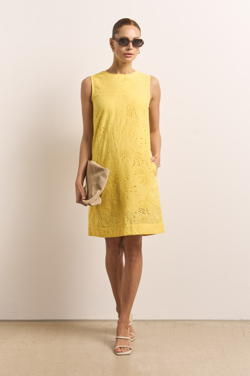Eine Frau posiert vor einer schlichten hellen Wand in einem gelben Baumwoll-Minikleid mit Ösen von Princess Goes Hollywood, beigen hochhackigen Sandalen und einer beigen Clutch. Ihr Haar ist zu einem ordentlichen Dutt gesteckt und sie trägt eine große runde Sonnenbrille für einen schicken Sommerlook.