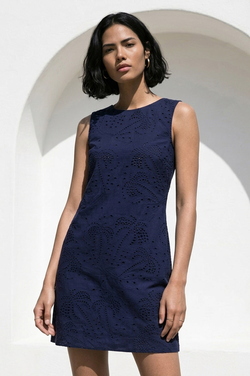 Eine Frau mit kurzen schwarzen Haaren trägt ein ärmelloses Baumwollminikleid mit Ösenstickerei von Princess Goes Hollywood. Sie steht vor einer weißen Wand mit einem gewölbten Ausschnitt und blickt selbstbewusst und mit neutralem Blick in die Kamera.