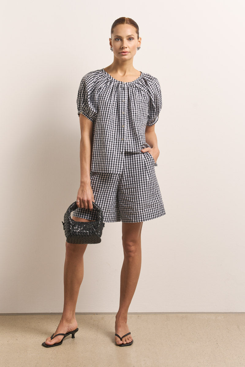 Eine Frau steht an einer schlichten Wand, trägt eine schwarz-weiße Vichy-Karo-Baumwollbluse mit Puffärmeln und dazu passende Shorts von Princess Goes Hollywood, hält eine kleine schwarze gewebte Handtasche in der Hand, trägt schwarze hochhackige Sandalen und hat ihr braunes Haar zurückgebunden.