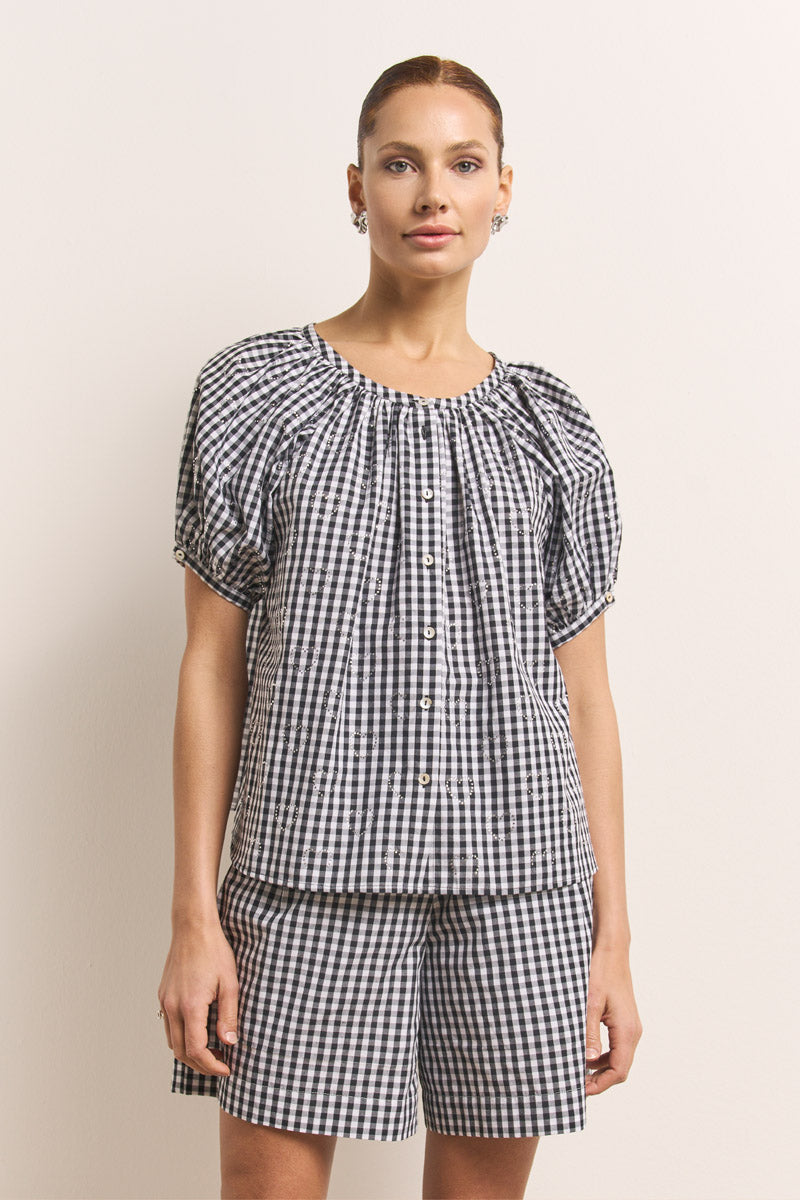 Eine Frau, die eine schwarz-weiße Vichy-Karo-Kurzarmbluse mit Puffärmeln und dazu passende Baumwollshorts von Princess Goes Hollywood trägt, steht vor einer schlichten hellen Wand. Ihr Haar ist zurückgebunden und sie trägt kleine Ohrringe.
