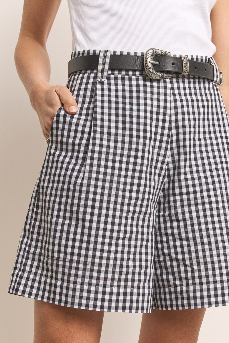 Eine Person trägt Princess Goes Hollywood schwarz-weiße Vichy-Karo Damen Shorts mit schwarzem Gürtel und silberner Schnalle, gepaart mit einem weißen ärmellosen Oberteil. Eine Hand steckt in der Tasche, nur der Unterkörper und der Oberschenkel sind sichtbar.