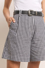 Eine Person trägt Princess Goes Hollywood schwarz-weiße Vichy-Karo Damen Shorts mit schwarzem Gürtel und silberner Schnalle, gepaart mit einem weißen ärmellosen Oberteil. Eine Hand steckt in der Tasche, nur der Unterkörper und der Oberschenkel sind sichtbar.