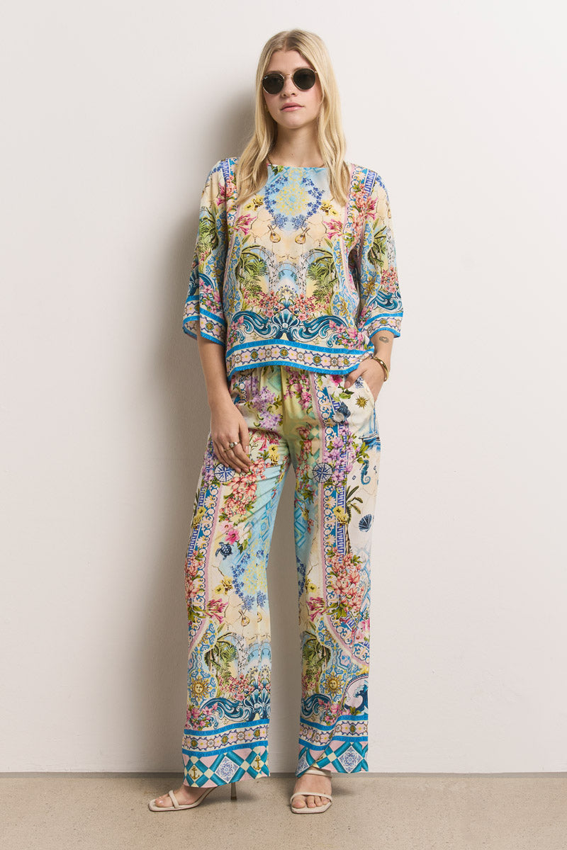 Eine Frau mit langen blonden Haaren und runder Sonnenbrille trägt eine lockere Princess Goes Hollywood Seidenbluse und eine Hose mit weitem Bein, die mit bunten, verschnörkelten Blumen- und Paisleymustern versehen ist. Sie steht mit Sandalen und den Händen in den Hosentaschen vor einer cremefarbenen Wand.