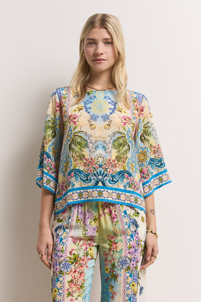 Eine Frau mit blondem Haar modelliert ein buntes, blumengemustertes Oberteil und eine passende Seidenhose mit elastischem Bund von Princess Goes Hollywood. Das blau-gelb-rosa-grüne Outfit ist mit verschnörkelten Mustern versehen, während sie vor einer weißen Wand steht.