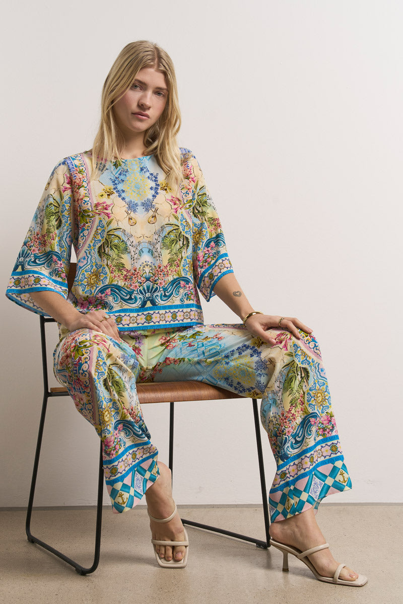 Eine Frau mit langen blonden Haaren sitzt auf einem Stuhl und trägt ein passendes Oberteil und eine Seidenhose von Princess Goes Hollywood mit floralen, geometrischen und landschaftlichen Motiven. Sie hat beigefarbene Absätze und steht vor einem einfachen Hintergrund mit hellen Wänden und Betonboden.