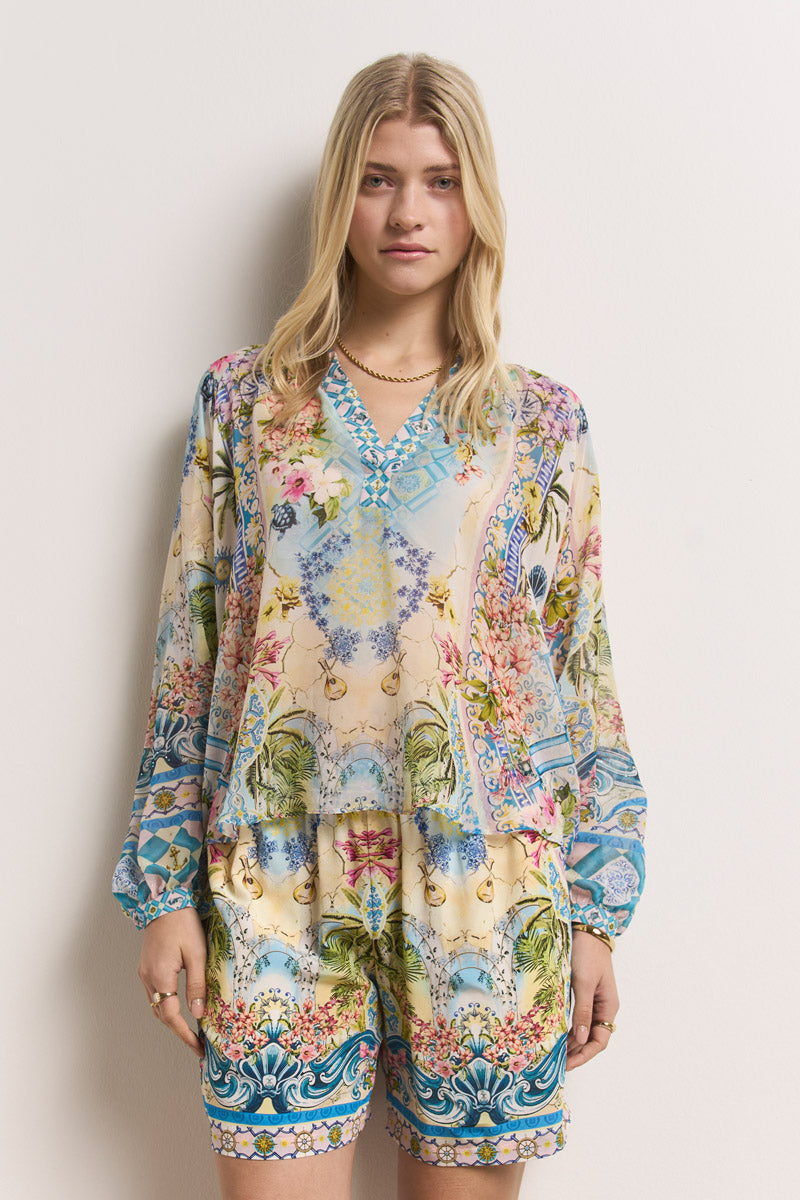 Eine blonde Frau steht vor einer weißen Wand und trägt einen langärmeligen Strampler von Princess Goes Hollywood mit einem lebhaften Blumen- und geometrischen Landschaftsmuster, einem V-Ausschnitt und weiten Ärmeln. Sie hat einen neutralen Gesichtsausdruck und trägt wenig Schmuck.