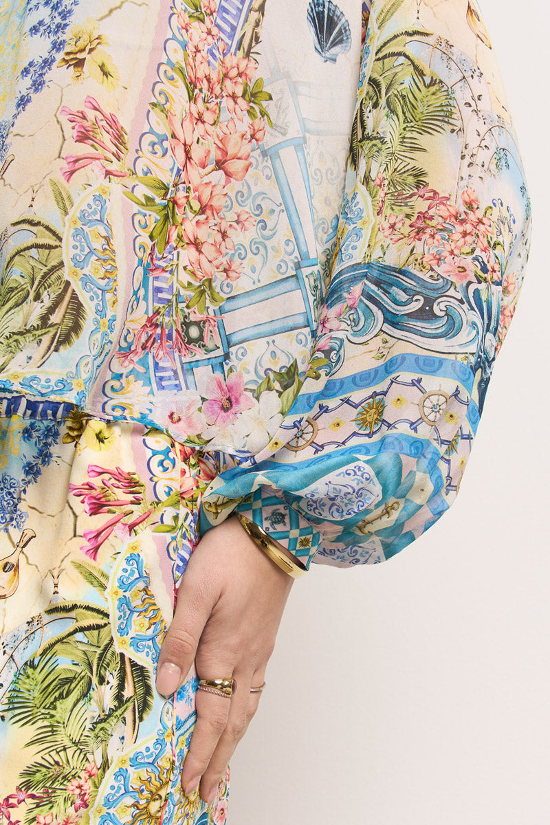 Eine Person trägt eine bunte, gemusterte Seidenbluse von Princess Goes Hollywood mit blauen, rosa und gelben Blumen- und Ornamentmustern. Die lockere, leicht durchsichtige Ballonärmel gibt den Blick auf goldene Ringe und einen Armreif frei; der Hintergrund ist schlicht hell.